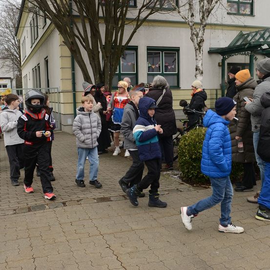 Eine Gruppe von Kindern und Erwachsenen in Kostümen geht um ein Gebäude mit Glasfenstern und einem grünen Dach herum.