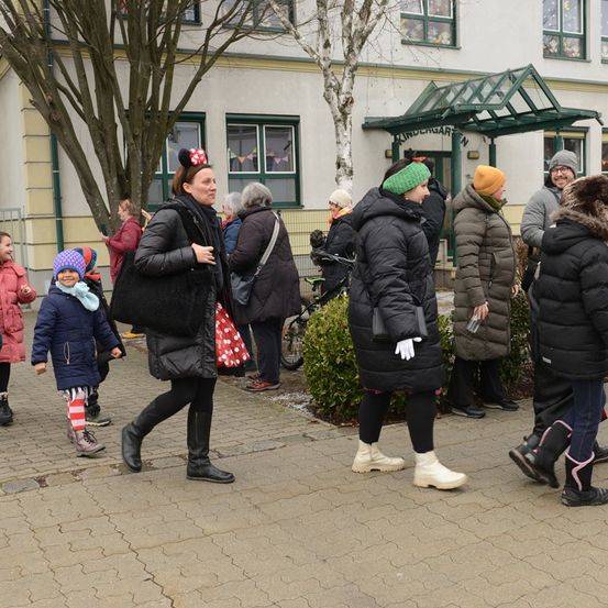 Menschen versammeln sich vor einem Gebäude, einige gehen, andere stehen. Eine Frau mit einer Minnie-Mouse-Kopfbedeckung geht mit einem Kind. Das Gebäude hat Glasfenster und einen grünen Vorbau.