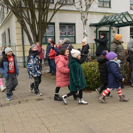 Eine Gruppe von Menschen, darunter Kinder und Erwachsene, geht in einer Reihe vor einem mit 'Kindergarten' beschrifteten Gebäude. Einige haben Mützen und Brillen. Das Wetter ist kalt, was an ihren Winterklamotten erkennbar ist.