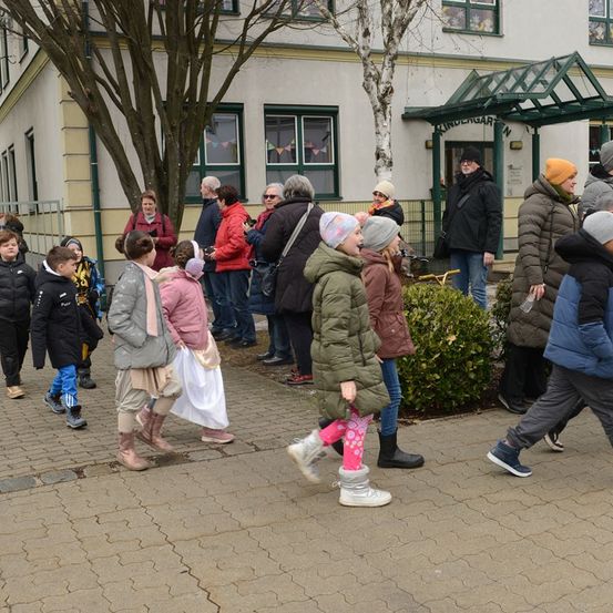 Eine Gruppe von Menschen geht auf einem gepflasterten Weg vor einem Gebäude. Sie tragen Winterkleidung. Einige tragen Mützen, Stiefel und Handschuhe.