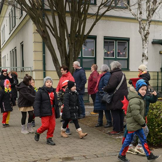Eine Gruppe von Kindern und Erwachsenen geht an einem Gebäude mit Bäumen vorbei, ein Kind trägt ein rotes Kostüm.
