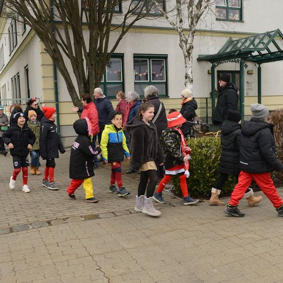 Eine Gruppe von Kindern in Winterkleidung geht auf einem gepflasterten Weg vor einem Gebäude mit grünem Dach.