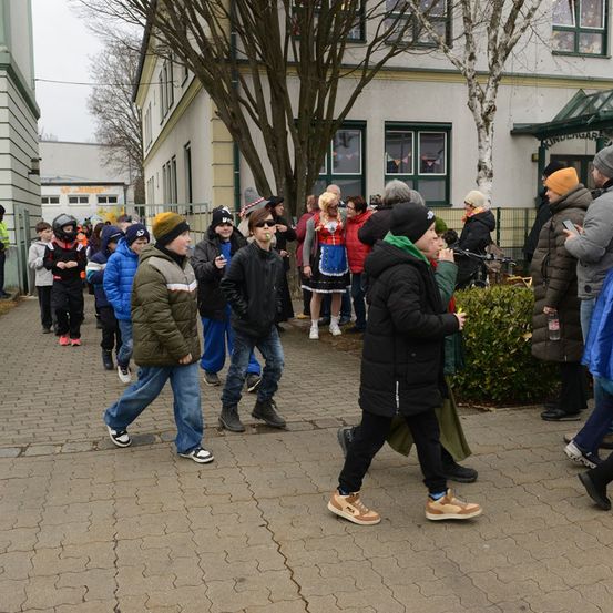 Eine Gruppe von Kindern in verschiedenen Winterkleidungen geht in einer Reihe auf einer Straße. Sie werden von Erwachsenen begleitet, von denen einige Fotos mit ihren Handys machen. Die Straße ist gepflastert.