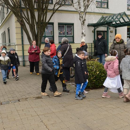 Eine Gruppe von Kindern und Erwachsenen steht vor einem Kindergarten. Einige tragen Wintermäntel, Mützen und Stiefel. Im Hintergrund sind Bäume und ein Gebäude zu sehen.