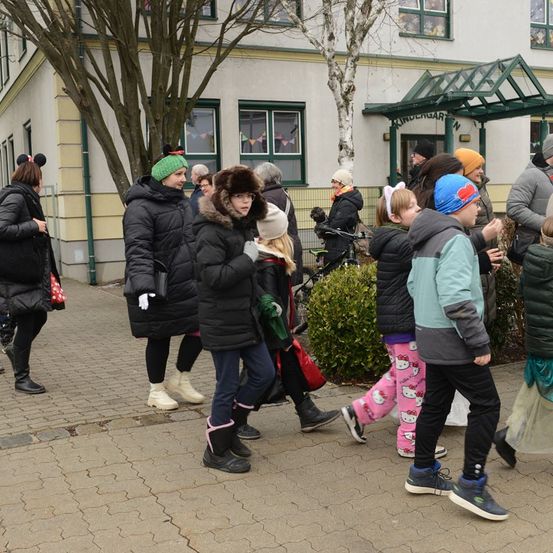 Mehrere Menschen gehen auf dem Bürgersteig vor einem Kindergarten. Der Kindergarten hat Glastüren und Fenster. Einige tragen Mützen, Stiefel und Handschuhe. Ein paar Fahrräder sind in der Nähe des Eingangs geparkt.