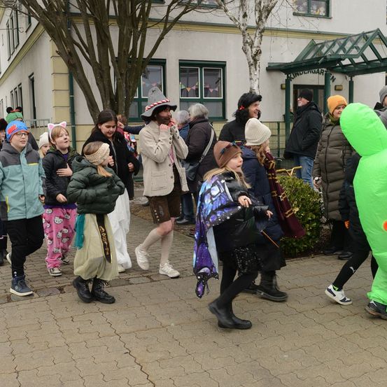 Eine Gruppe von Kindern und Erwachsenen, einige in Kostümen, stehen vor einem Gebäude mit Glasfenstern und einem Glaseingang. Die Gruppe umfasst eine Person in einem grünen aufblasbaren Kostüm.
