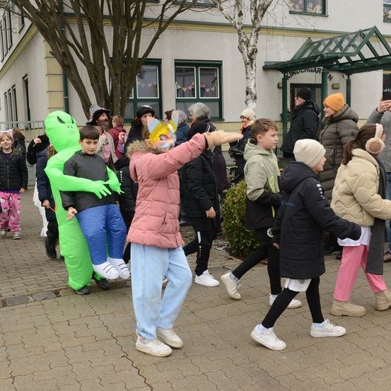 Eine Gruppe von Kindern und Erwachsenen ist in Kostümen gekleidet vor einem Gebäude. Einige tragen Masken und eine Person zeigt. Das Wetter ist kalt und die Leute tragen Winterkleidung.