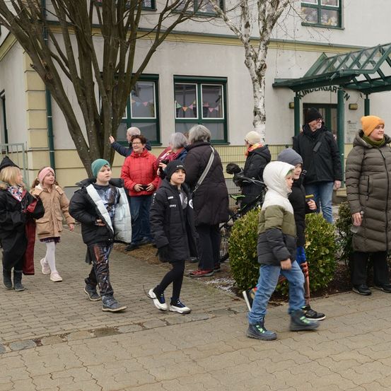 Eine Gruppe von Kindern und Erwachsenen in Winterkleidung geht vor einem Gebäude mit einem Schild namens Kindergarten. Einige tragen Mützen und einige haben Taschen bei sich. Vor dem Gebäude stehen Bäume.