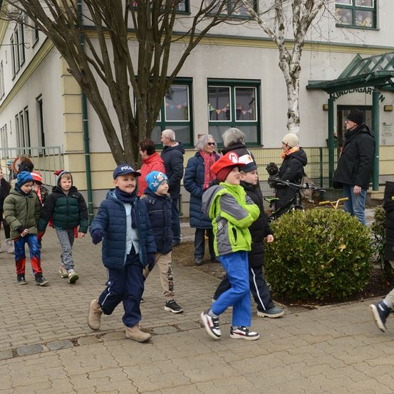 Mehrere Kinder und Erwachsene gehen in einer Reihe vor einem Gebäude. Ein Kind trägt einen Mario-Hut.