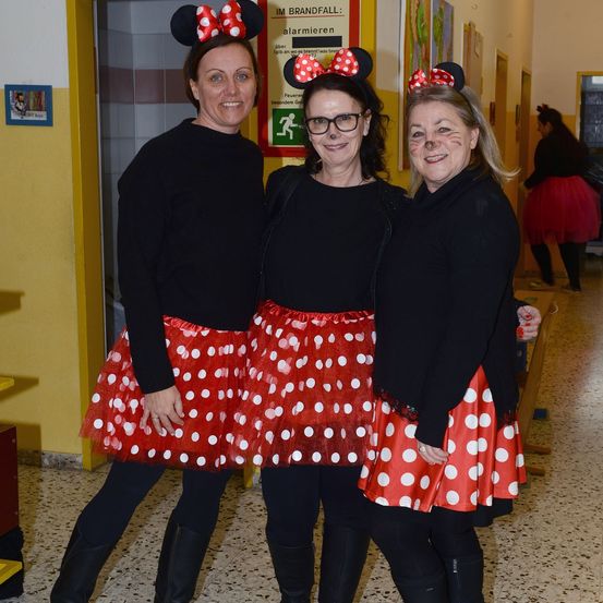 Drei Frauen in Minnie-Mouse-Kostümen, die lächelnd für ein Foto in einem Flur posieren. Die Frau auf der linken Seite trägt eine Minnie-Mouse-Kopfband und Stiefel.