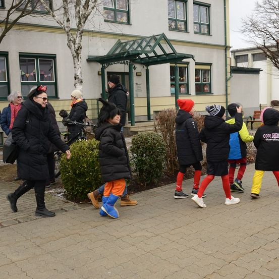 Eine Gruppe von Kindern in Winterkleidung geht auf dem Bürgersteig vor einem Gebäude. Einige tragen Mützen und Stiefel.