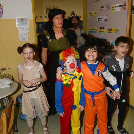 Eine Gruppe von Kindern und ein Erwachsener, der als Clown verkleidet ist, stehen gemeinsam in einem Raum mit gelber Wand.