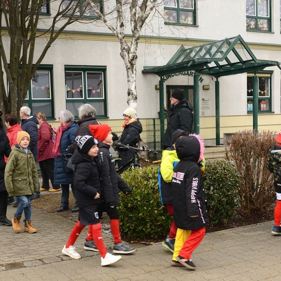 Eine Gruppe von Kindern und Erwachsenen versammelt sich vor einem Kindergarten, einige in Winterkleidung und Mützen. Im Hintergrund sind Bäume und ein Gebäude mit Glasfenstern zu sehen.