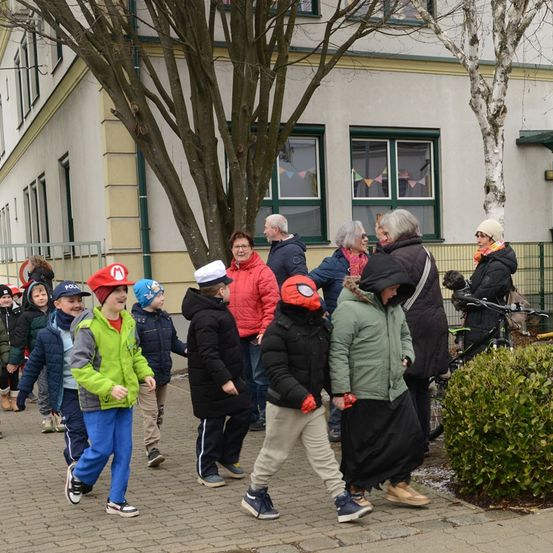 Eine Gruppe von Kindern und Erwachsenen, einige mit Mützen, gehen vor einem Gebäude. Sie sind für kaltes Wetter gekleidet. Bäume und Pflanzen umgeben das Gebiet.