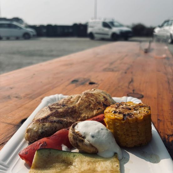 Ein Teller mit gegrilltem Hähnchen, Mais und Gemüse auf einem Holztisch mit geparkten Autos im Hintergrund.