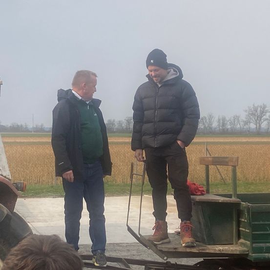 Zwei Männer stehen auf einem Anhänger in einem Feld. Einer trägt eine schwarze Jacke und Hose, der andere ein grünes Shirt. Im Hintergrund sind ein Feld mit Pflanzen und Bäumen zu sehen.