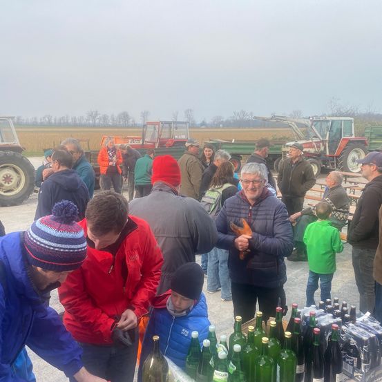 Eine Gruppe von Menschen steht um einen Tisch mit Flaschen herum. Einige tragen Winterkleidung. Dahinter sind Traktoren und Maschinen geparkt. Das Gelände ist von Feldern und Bäumen umgeben.