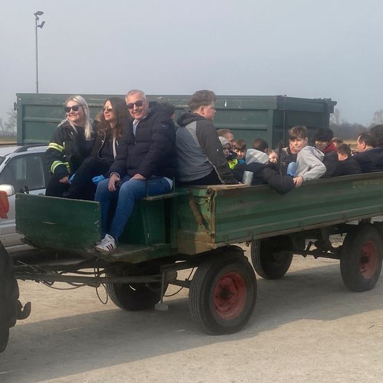 Eine Gruppe von Erwachsenen und Kindern fährt in einem grünen Anhänger, der von einem Fahrzeug gezogen wird, mit grünen Containern und einem Auto im Hintergrund.