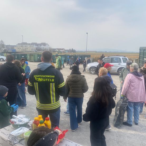 Eine Gruppe von Menschen versammelt sich um einen Tisch mit Essen. Ein Mann in einer Feuerwehruniform steht in der Mitte. Im Hintergrund befinden sich Autos und Müllcontainer.
