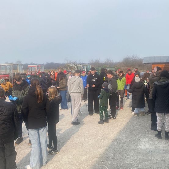 Eine Gruppe von Menschen, darunter Kinder, steht im Freien auf einer Kiesfläche, einige tragen Handschuhe. Im Hintergrund sind Maschinenfahrzeuge geparkt.