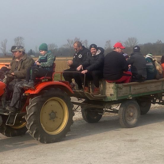 Mehrere Personen sitzen auf einem Traktor mit Anhänger auf einer Straße, ein junger Mensch in einer grünen Jacke sitzt vorne.