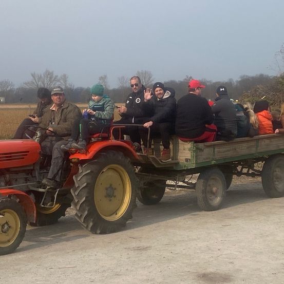 Eine Gruppe von Menschen fährt auf einem roten Traktor mit Anhänger auf einer Landstraße. Sie tragen Winterkleidung und einige benutzen ihre Telefone.