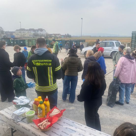 Eine Gruppe von Menschen, darunter Kinder, versammelt sich um einen Tisch mit Essen und Getränken. Ein Feuerwehrmann steht in der Nähe. Im Hintergrund befinden sich Müllcontainer und ein Auto.