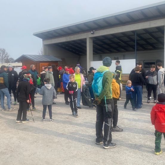 Eine Gruppe von Menschen steht in einer Reihe in einem Außenbereich. Einige von ihnen tragen Hüte und Brillen. Einige halten Taschen und andere tragen Rucksäcke.