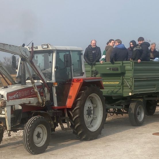 Ein Traktor mit einer großen Gruppe von Menschen, die auf einem Anhänger sitzen, der an den Traktor angeschlossen ist. Der Traktor ist weiß und rot mit großen Reifen.