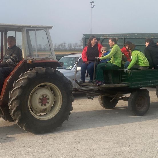 Ein älterer Mann fährt einen Traktor mit Anhänger, auf dem mehrere Personen sitzen. Sie tragen grüne Mäntel.