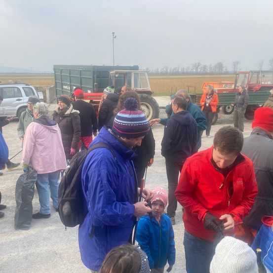 Eine Gruppe von Menschen versammelt sich um einen Mann, wahrscheinlich auf einem Feld. Traktoren und ein Lkw sind im Hintergrund. Der Mann hält etwas für einen jungen Jungen mit einer rosa Mütze.