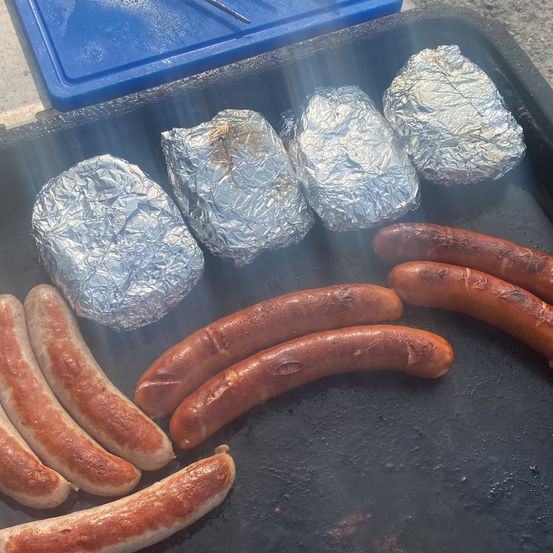 Ein Grill mit mehreren Würstchen und eingewickelten Lebensmitteln, möglicherweise Kartoffeln, die auf einer schwarzen Oberfläche kochen.