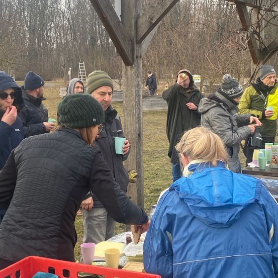 Eine Gruppe von Menschen versammelt sich um einen Tisch auf einem Feld, trägt Winterkleidung und hält Tassen. Eine Person lächelt, während eine andere trinkt. Im Hintergrund sind Bäume und eine Leiter zu sehen.