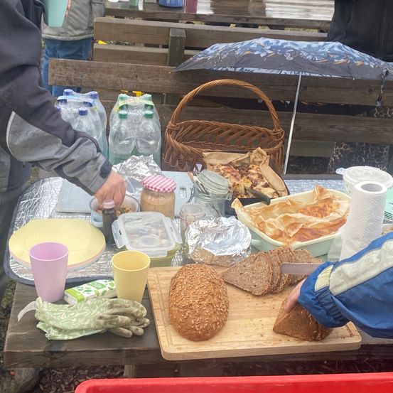Ein Tisch im Freien hat ein Schneidebrett mit Brot, einem Korb und einer Flasche Marmelade. Eine Person schneidet das Brot, und eine andere Person steht in der Nähe. Wasserflaschen, Teller und andere Gegenstände sind auf dem Tisch.