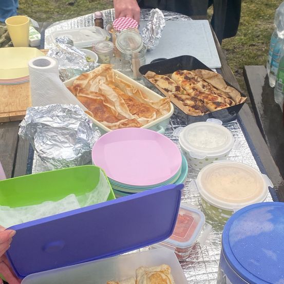 Ein Outdoor-Tisch mit verschiedenen Lebensmitteln, einschließlich eingewickelter Lebensmittel, Schalen und Behältern. Es gibt auch Tassen, Flaschen und ein Schneidebrett. Der Tisch ist mit Folie bedeckt.