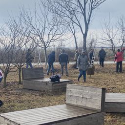 A group of people dressed in warm clothing work in an outdoor area, some standing by wooden structures, others by trees. A cloudy sky is visible above.