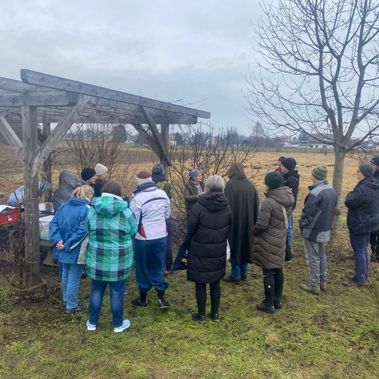 Eine Gruppe von Menschen in warmer Kleidung steht in der Nähe einer Holzstruktur und unterhält sich. Im Hintergrund befinden sich Bäume, Büsche und ein Gebäude in der Ferne unter einem bewölkten Himmel.