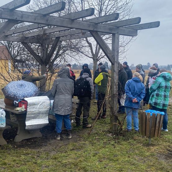 Eine Gruppe von Menschen in Winterkleidung steht unter einem Holzpavillon auf einem Rasen. Einige schauen auf einen Picknicktisch mit einem Regenschirm und anderen Gegenständen.