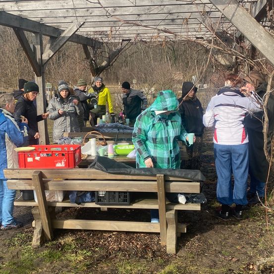 Eine Gruppe von Menschen versammelt sich unter einer Holzstruktur, möglicherweise in einem Wald, mit Tischen, Körben und Lebensmitteln. Einige tragen Jacken und Mützen.