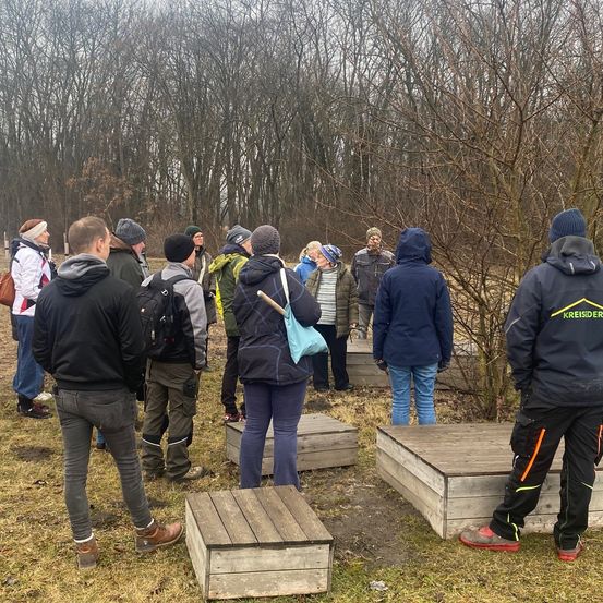 Eine Gruppe von Menschen steht in einem Grasfeld und trägt Winterkleidung. Sie scheinen auf einem Ausflug zu sein, mit Holzkisten in der Nähe. Eine Frau trägt eine blaue Tasche.