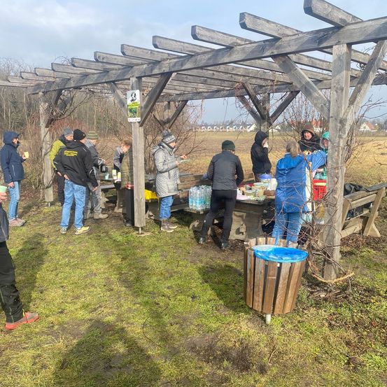 Eine Gruppe von Menschen versammelt sich unter einem Holzpavillon. Sie beschäftigen sich mit Aktivitäten, einige stehen, andere sitzen. Flaschen und ein Mülleimer sind vorhanden. Der Ort befindet sich in einem Grasfeld mit Bäumen im Hintergrund.