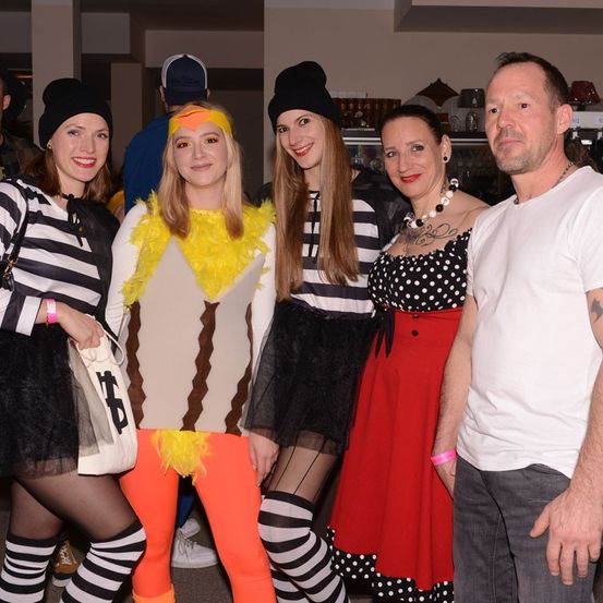 Eine Gruppe von Menschen in Kostümen, zwei mit Hühnerkostümen, eine mit einem gepunkteten Kleid und ein Mann in einem weißen T-Shirt, posiert für ein Foto.