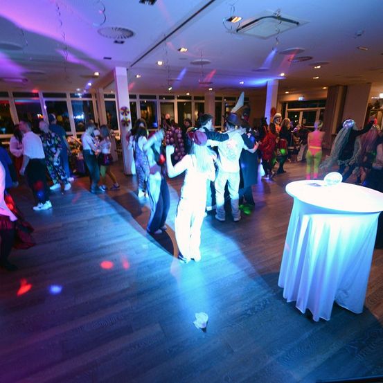 Eine Gruppe von Menschen tanzt auf einem Holzboden in einem Partysaal mit Disko-Lichtern. Die Lichter spiegeln sich auf dem Boden. Ein Tisch mit einem weißen Tuch steht rechts.