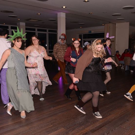 Eine Gruppe von Erwachsenen tanzt auf einer Party in einem Raum mit Holzboden, einige in Kostümen.