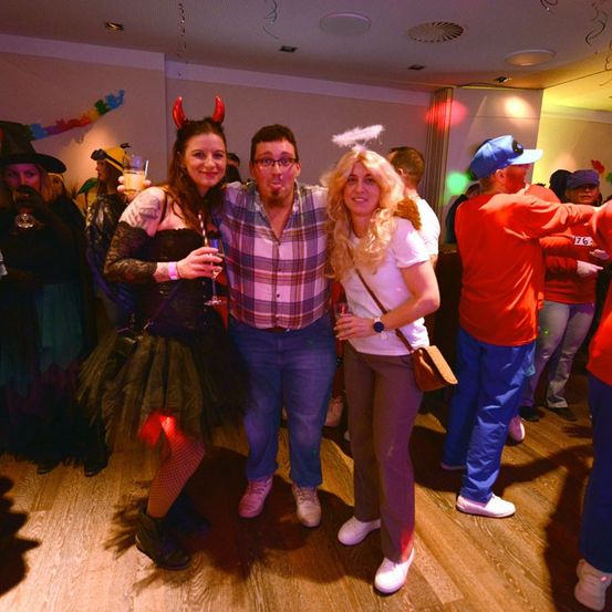 Drei Menschen in Kostümen stehen in einem Partysaal. Eine Frau in einer Hexen-Kostüm, ein Mann in einem karierten Hemd und eine Frau mit blonden Haaren. Andere Menschen sind im Hintergrund, und der Raum ist dekoriert.