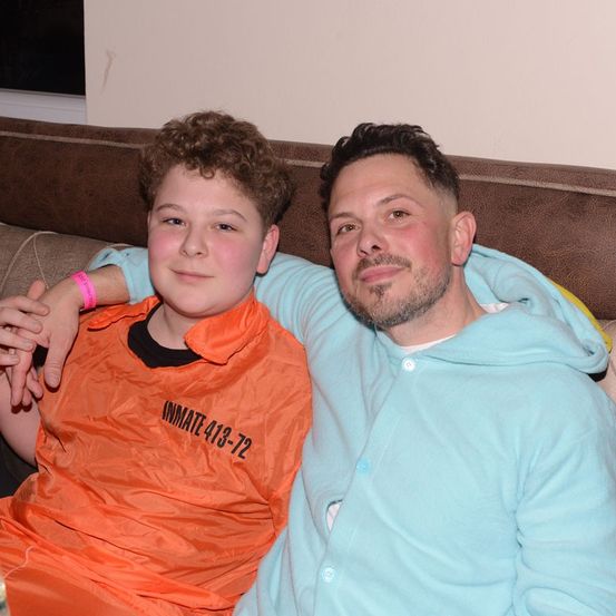 Ein Mann und ein Junge sitzen gemeinsam auf einem Sofa, der Junge in einem orangefarbenen Häftlingsoverall und der Mann in einer blauen Kapuzenjacke. Der Junge lächelt und lehnt seinen Kopf an die Schulter des Mannes.