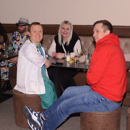 Eine Gruppe von Menschen sitzt um einen Tisch in einem Restaurant. Eine Person trägt einen Laborkittel und ein Stethoskop. Eine andere Person trägt eine rote Kapuzenjacke. Der Tisch ist mit Weingläsern, Bechern und anderen Gegenständen gedeckt.