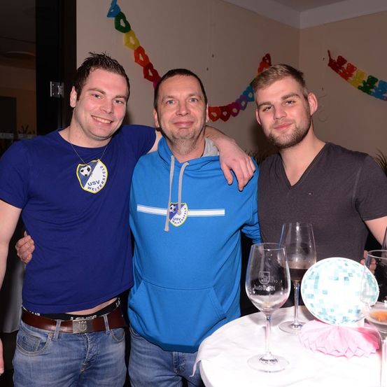 Drei Männer stehen in einem Raum mit einem Tisch vor ihnen. Sie lächeln und scheinen für ein Foto zu posieren. Der Mann links trägt einen Gürtel und eine Halskette. Der Mann in der Mitte trägt eine blaue Kapuzenjacke. Der Mann rechts trägt ein schwarzes T-Shirt. Auf dem Tisch stehen Weingläser, eine Discokugel und eine rosa Serviette.