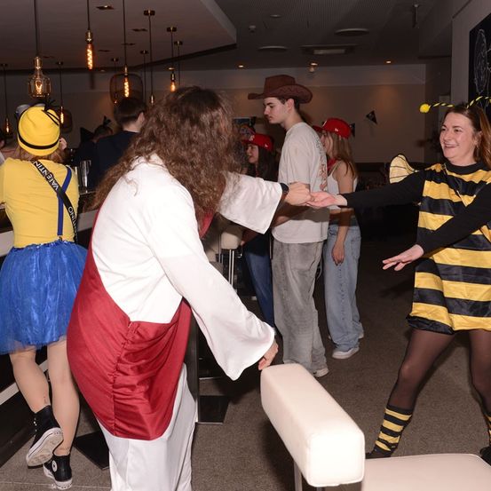 Menschen in Kostümen tanzen in einem Club. Eine Frau in einem Bienenkostüm schüttelt einem Mann in einem Gewand die Hand.