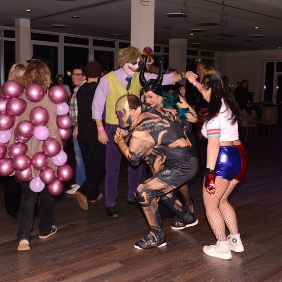 Eine Gruppe von Menschen in Kostümen tanzt in einem Saal mit Ballons, einer Säule und Stühlen.